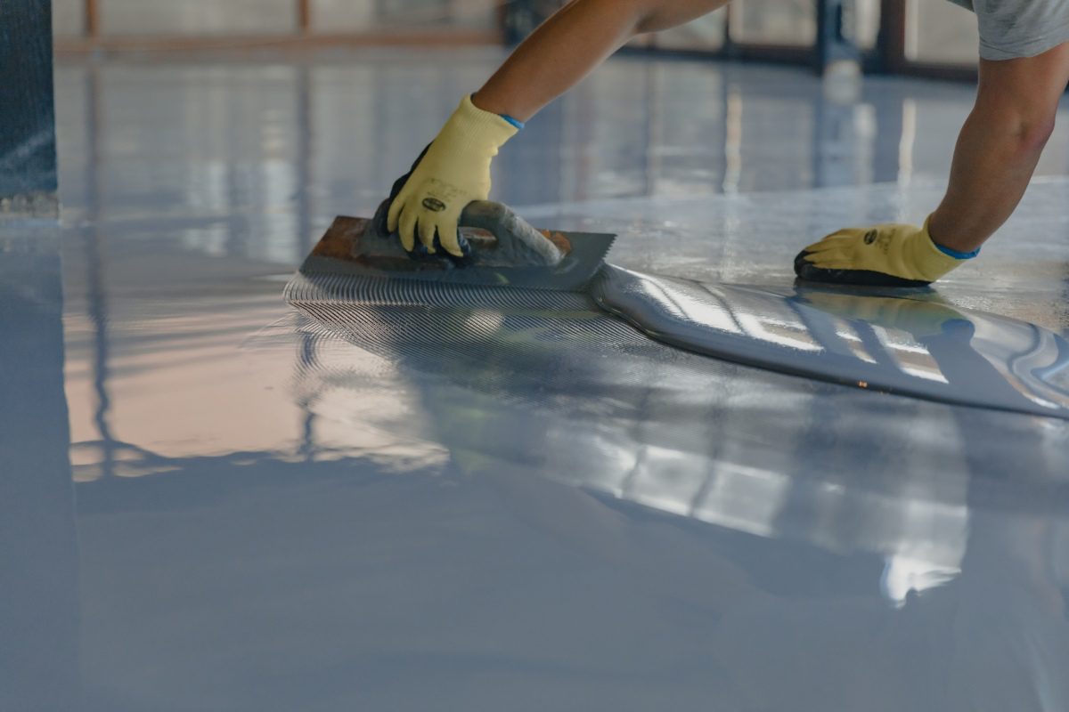 A person wearing gloves is smoothing a liquid epoxy floor with a trowel, creating a glossy surface.