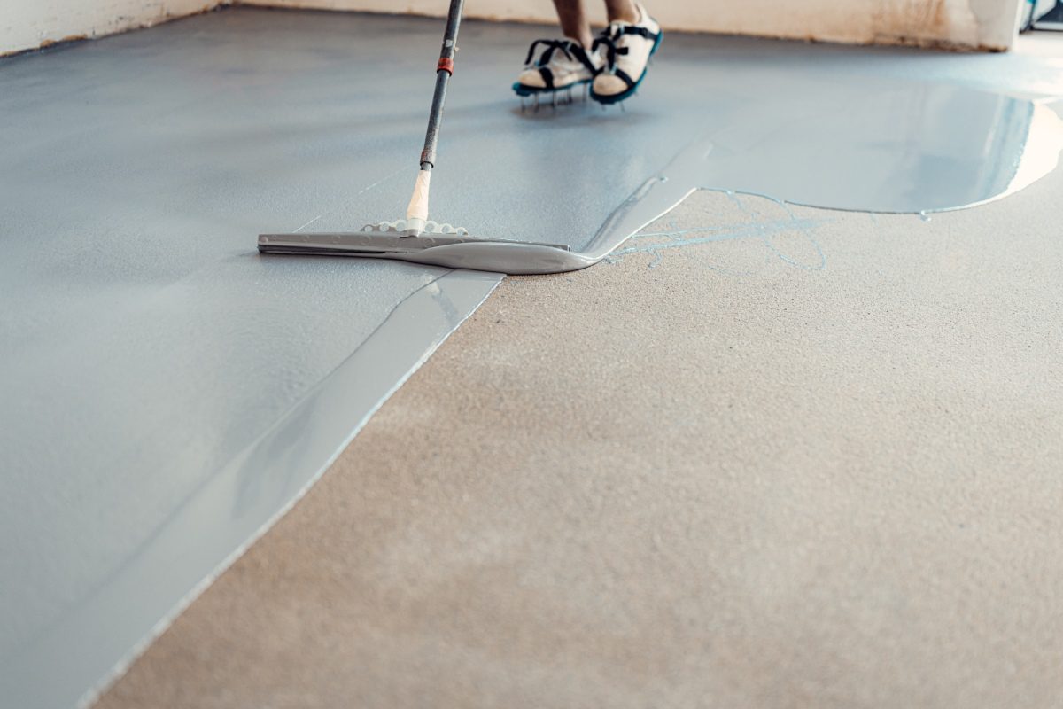 A person uses a squeegee to spread gray epoxy coating on a concrete floor. Their feet are equipped with spiked shoes.