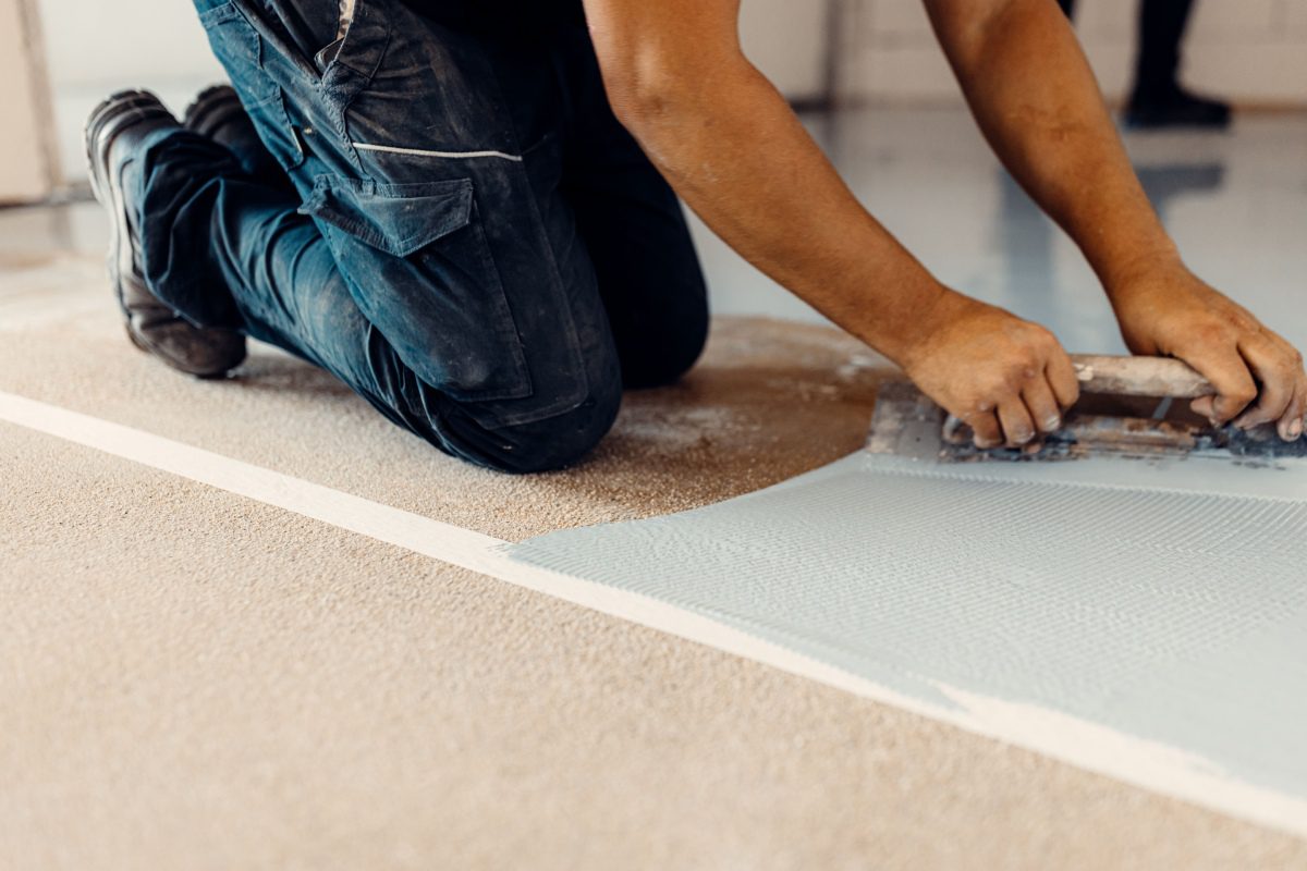 Person kneeling on the floor, using a trowel to apply a light-colored material onto a textured surface.